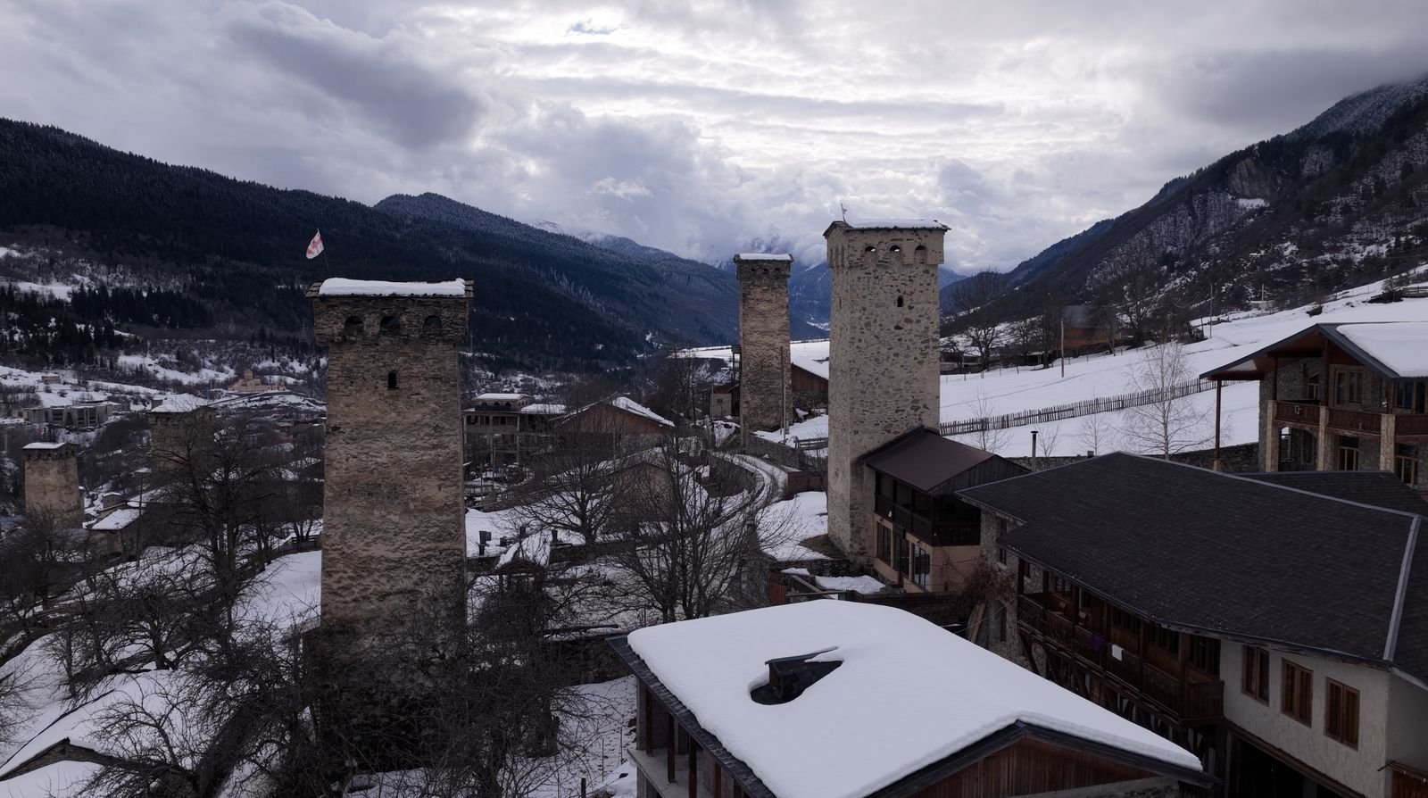 Medieval stone towers of Mestia with a Georgian flag above snow-covered roofs and the winter valley