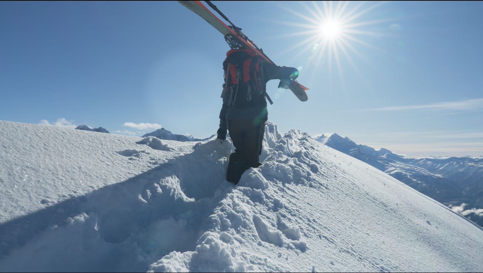 Skier boot-packing along a snowy ridge with skis on his shoulder, facing the winter sun